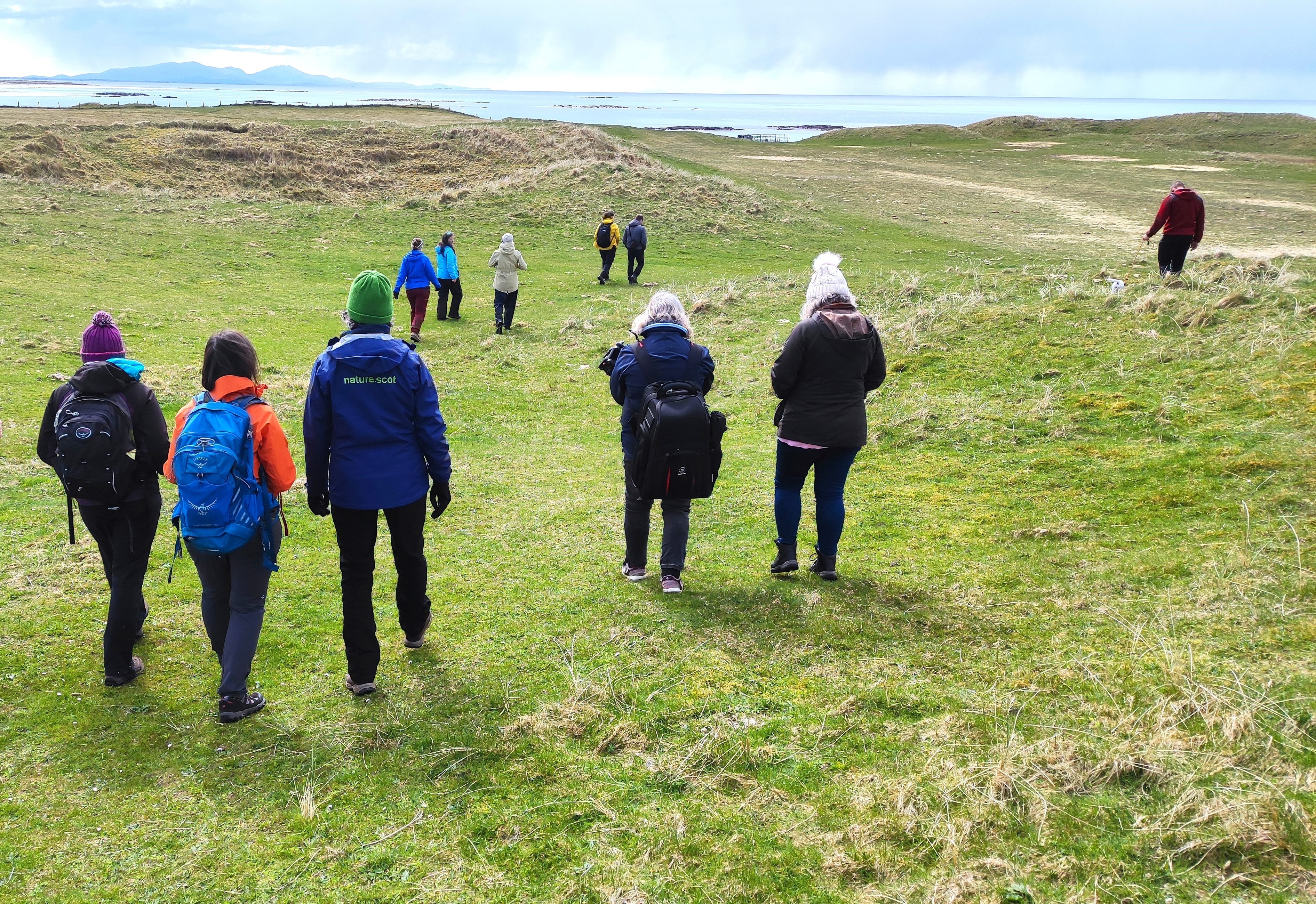 A group of people walk towards the shore through grassy dunes