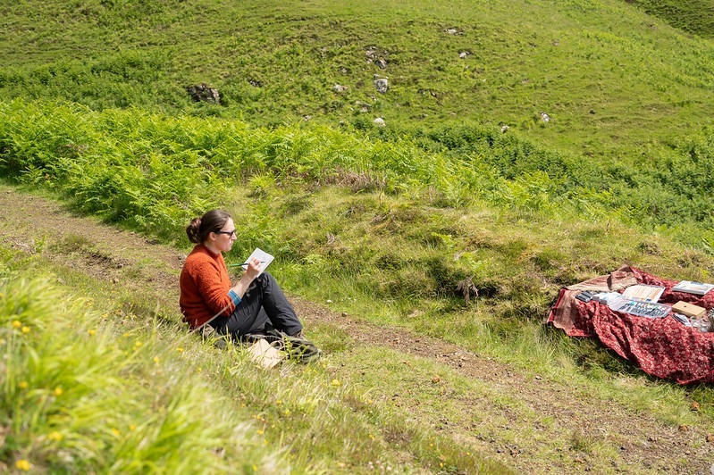 A person sits on grass and holds a notebad and pen