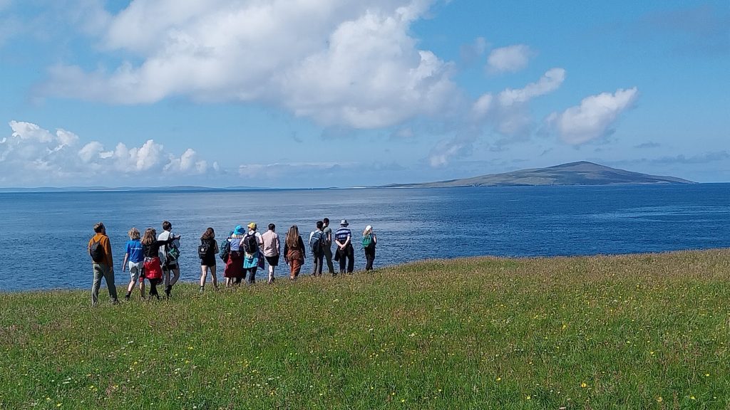 A group of people stand on a grassy hilltop. Beyond them is the sea.