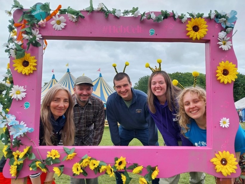 A groupd of people peer through a lrage pink frame adorned with flowers. Two wear bee antennae headbands on their heads.