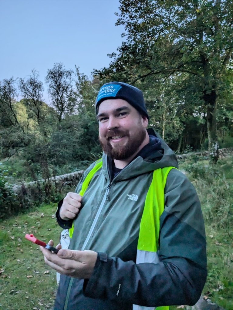 A man holds a bat detector. He is wearing a hat with the Species on the Edge logo on it and a high vis vest