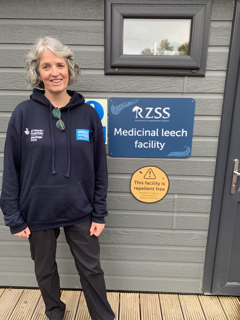 The Species in the Edge Programme Manager, Fiona, stands beside a sign on the side of a building which reads 'RZSS Medicinal leech facility'