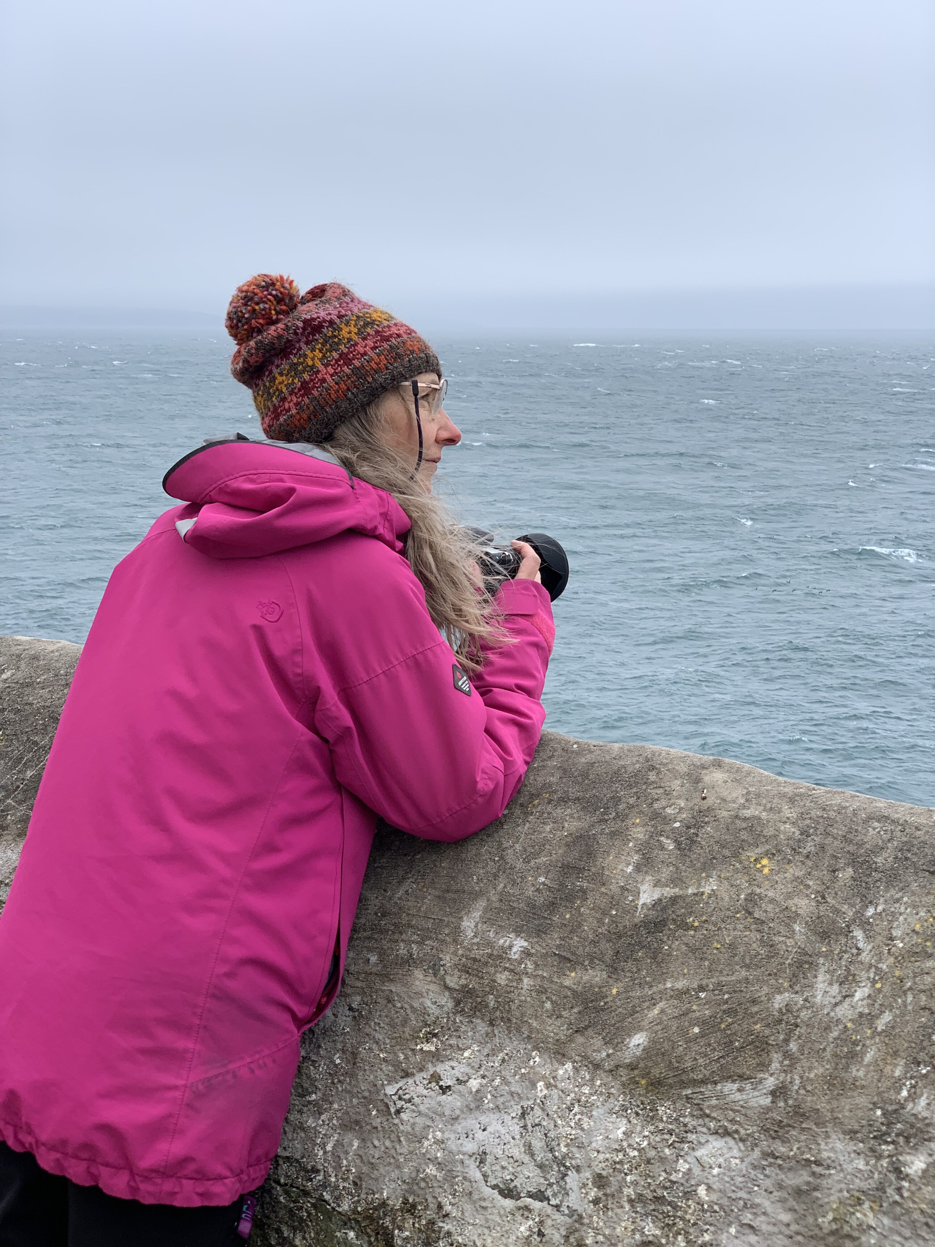 A woman leans on a wall and looks out to sea. She is holding a camera.