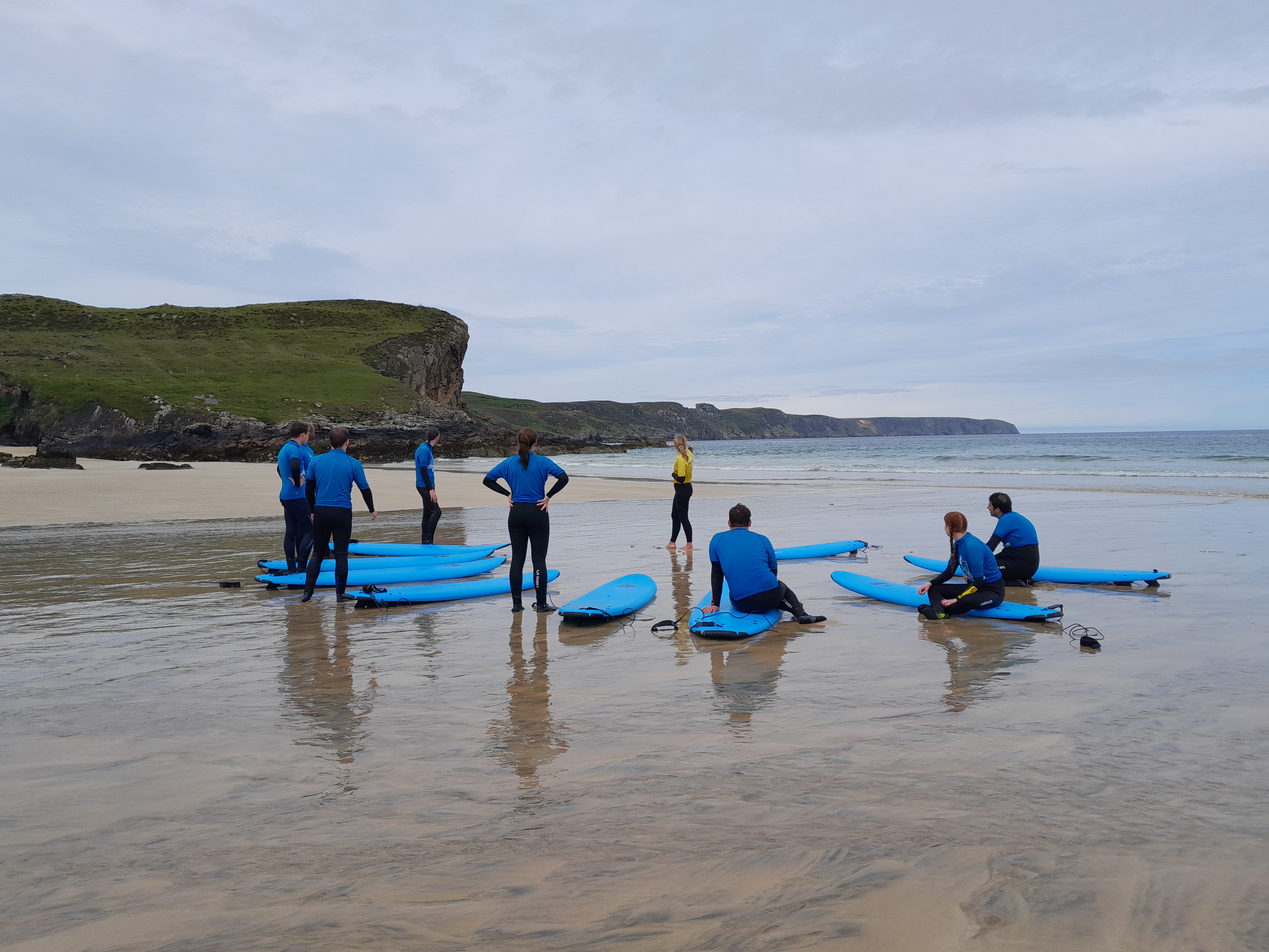 A group of people on a beach with surf boords. Behind them is a dramatic coast line of cliffs.