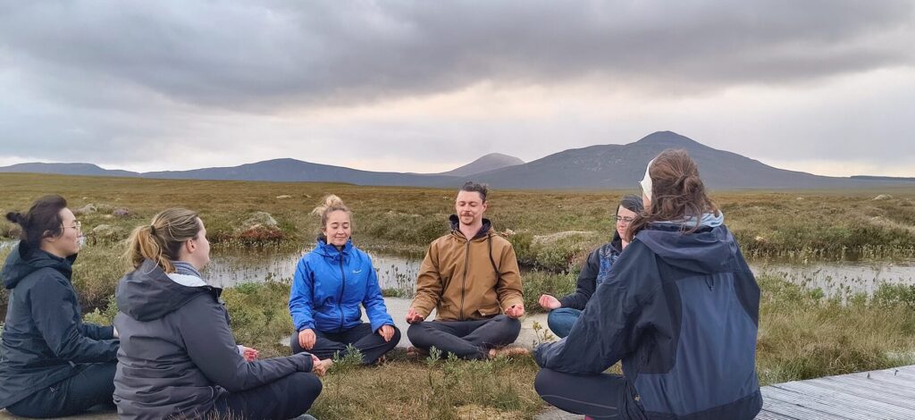 A group of of people sit outside meditating. Mountains can be seen in the background