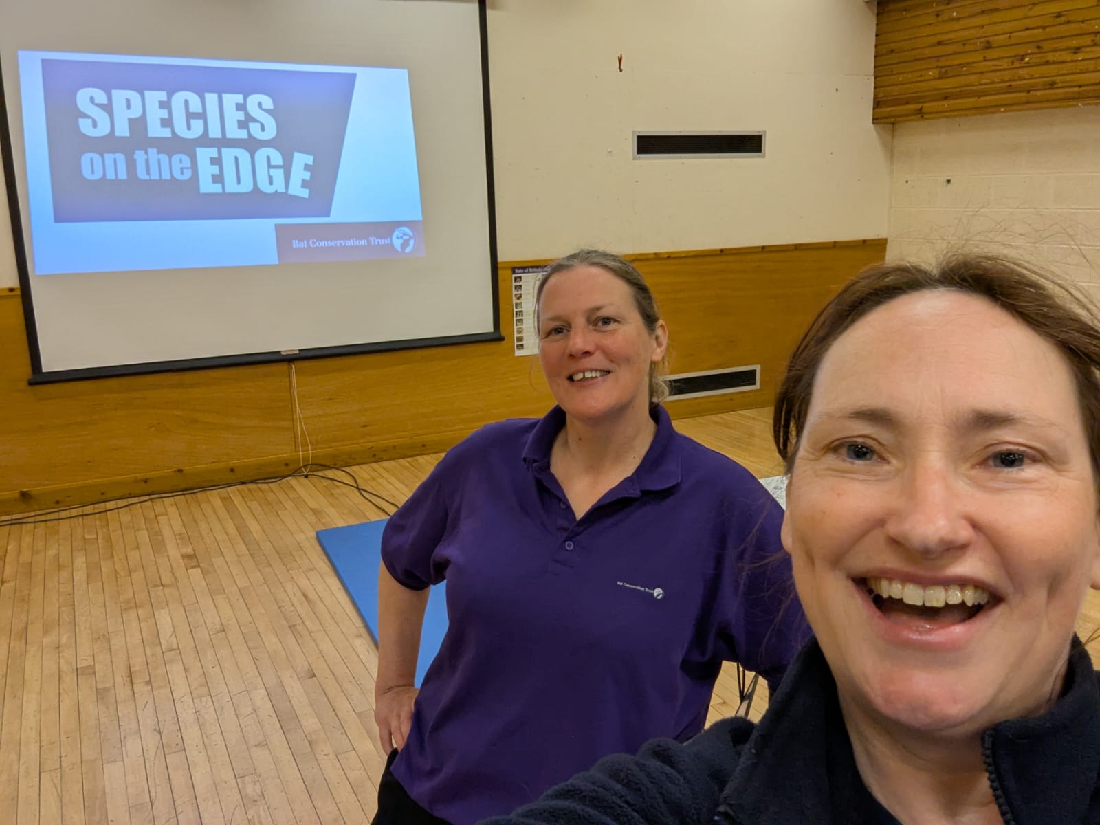 Two people take a selfie in a school hall in front of a projection of the Species on the Edge and Bat Conservation Trust logos.