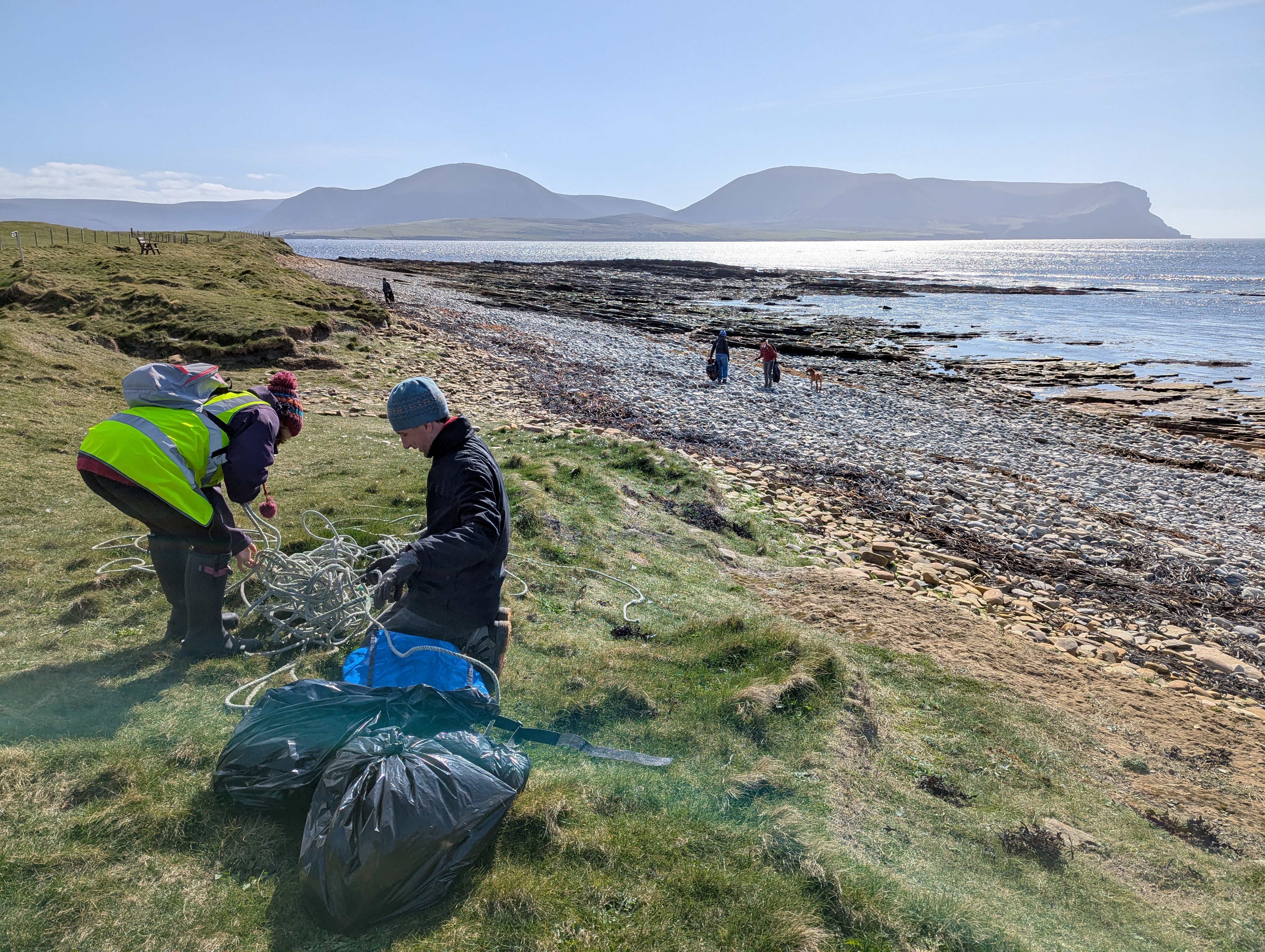 A photo of people clearing rubbish on a pebbly beach in Orkney. In the foreground is two people clearing rope from a grassy banking. They have black rubbish bags in front of them. One person is kneeling and the other, wearing a high vis vest, is standing bent over. In the middle ground is a pebbly beach and three people can be seen on the beach in the distance. In the background is a glittering sea, a land mass and a blue sky.