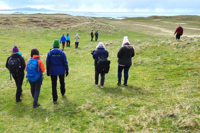 A group of people walk towards the shore through grassy dunes