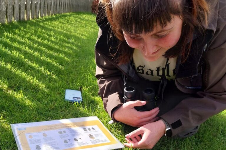 A person with brown hair and a fringe is knelt own on the grass, bent over, looking at a bee in the grass. Lying beside the person on the grass is a bumblebee information sheet. The person in smiling.