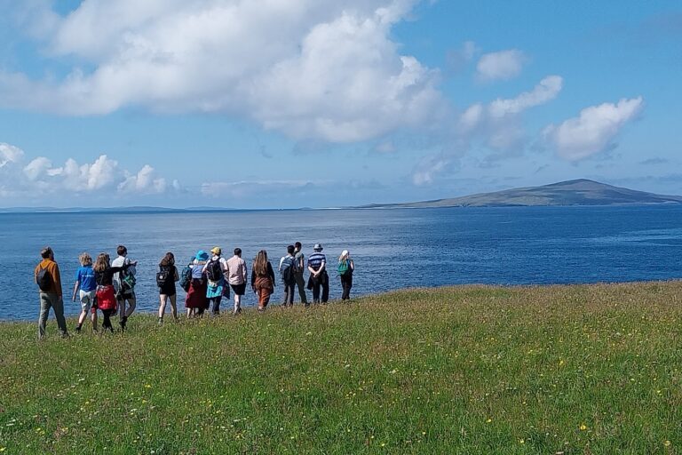 A group of people stand on a grassy hilltop. Beyond them is the sea.