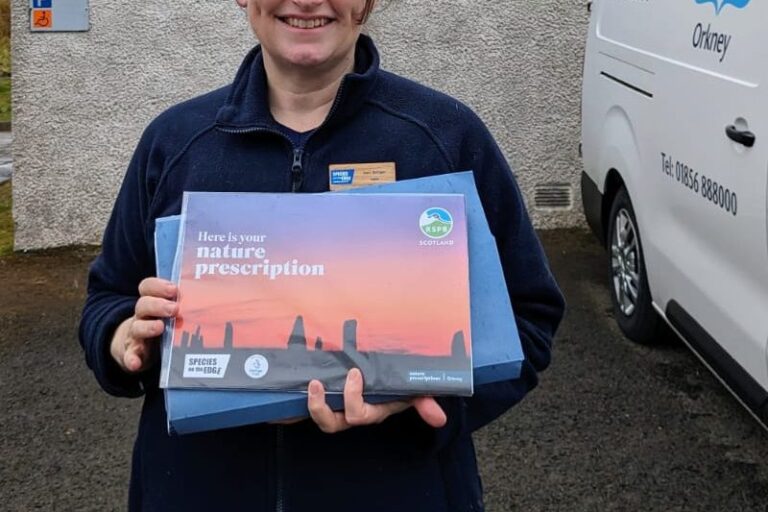 A woman stands in front of a GP surgery in Orkney. She holds a Nature Presciprion Calendar. The calendar has a bright sunset over the Ring of Brodgar on the front.