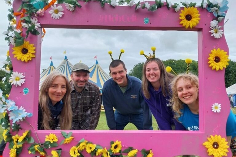 A groupd of people peer through a lrage pink frame adorned with flowers. Two wear bee antennae headbands on their heads.