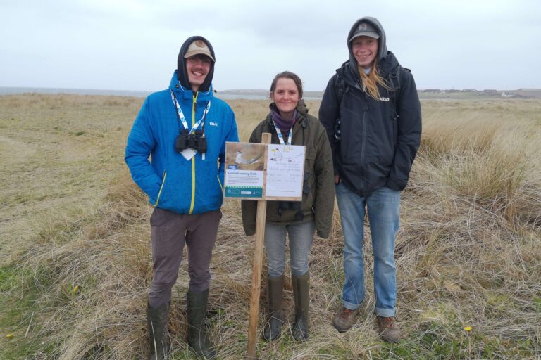 Three people stand behind a sign with a picture of a tern on it. The sign provides information on tern disturbance.