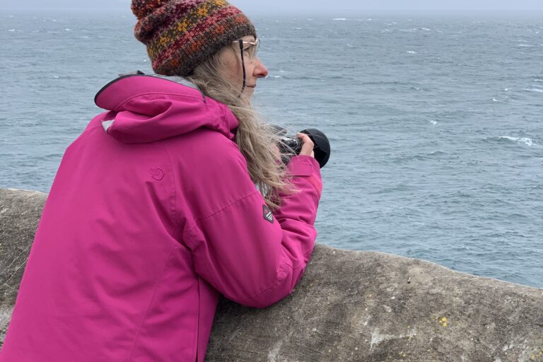 A woman leans on a wall and looks out to sea. She is holding a camera.