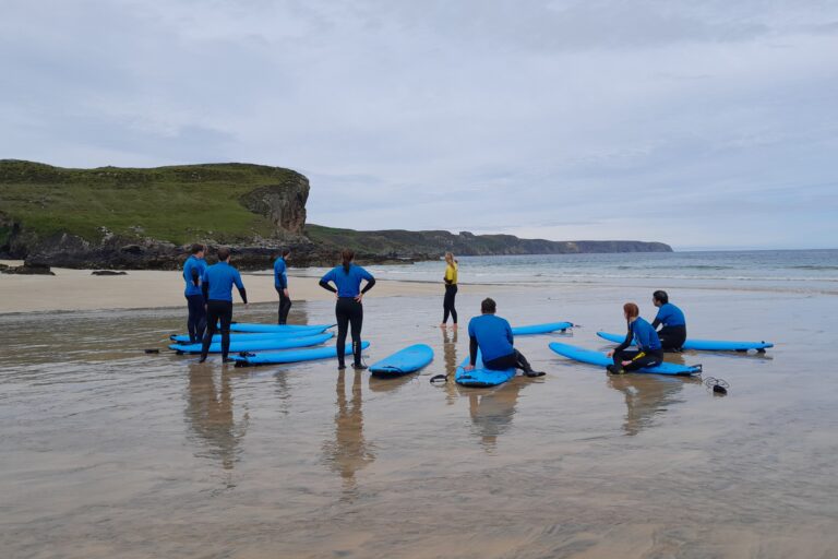 A group of people on a beach with surf boords. Behind them is a dramatic coast line of cliffs.