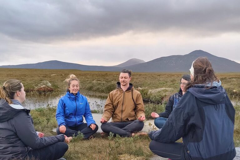 A group of of people sit outside meditating. Mountains can be seen in the background